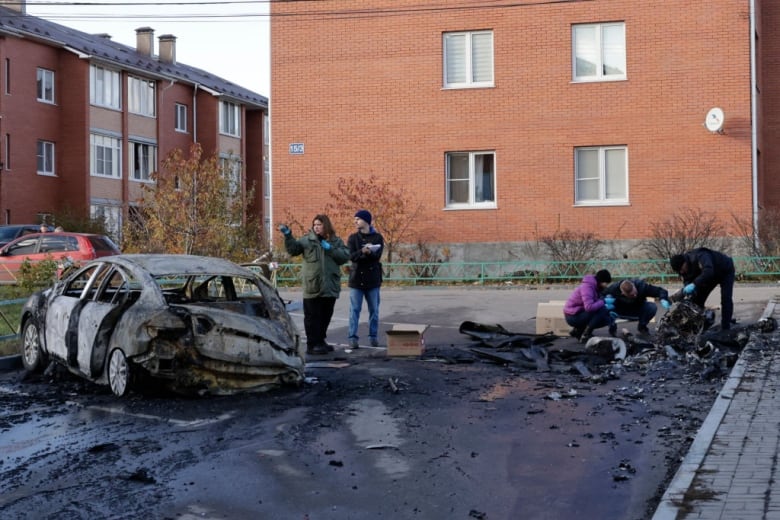 People gather near the wreckage of a burnt-out car. 