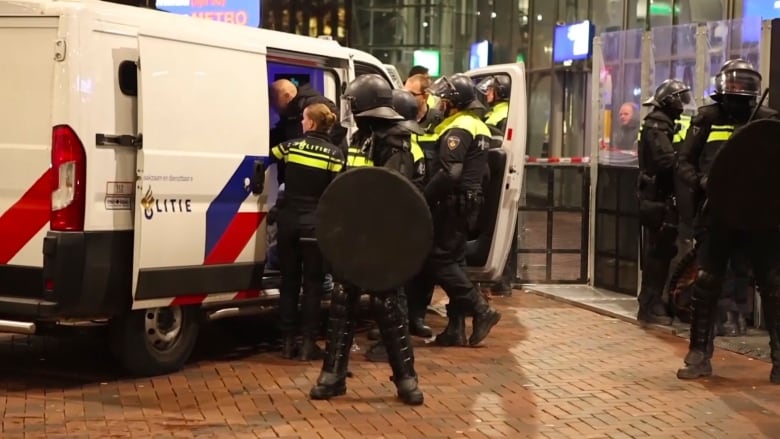 A group of police officers wearing riot gear stand next to a police van.