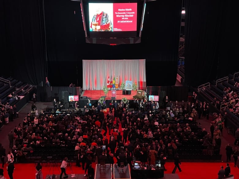 A crowd in a room in front of a stage with a portrait, flags, flowers.