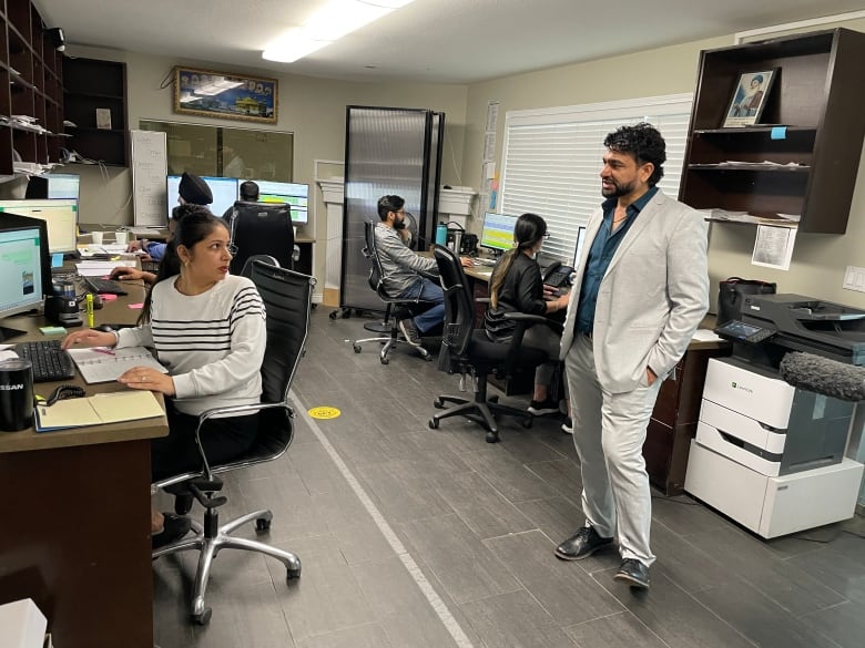 A man in a light-coloured suit talks to a woman as she works at a desk in a small office with several other people working at nearby computers. 