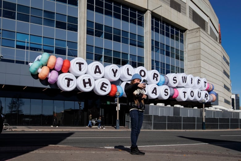 A woman takes a selfie in front of Rogers Centre stadium where hangs a giant friendship bracelet sign that reads: Taylor Swift The Eras Tour.