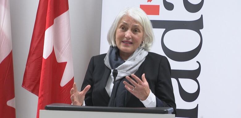 A woman with gray hair speaks in front of a podium. Behind her are Canadian flags and a big whiteboard with the word Canada written on it.