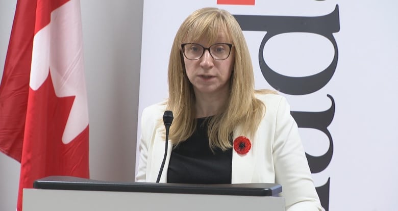 A woman wearing glasses speaks in front of a podium. Behind her is a Canadian flag.