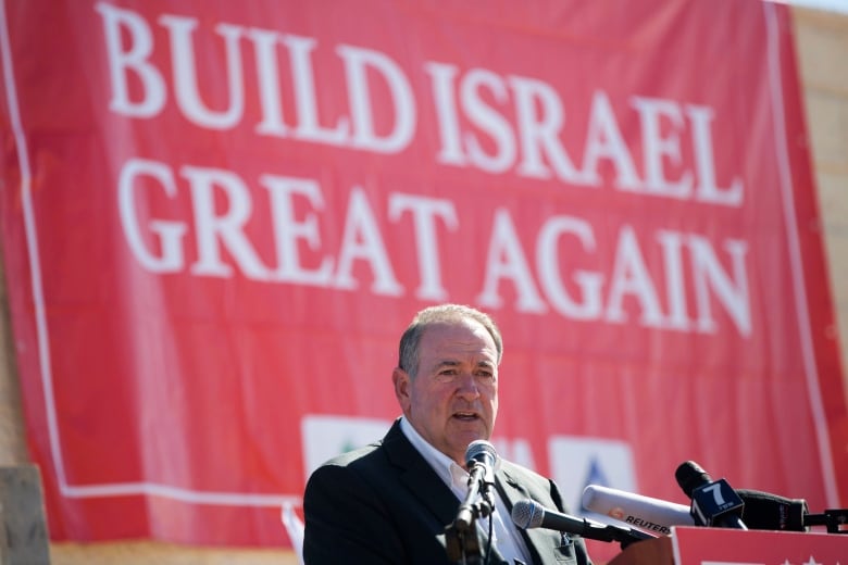 A man in a suit stands at a podium. A red banner behind him says 