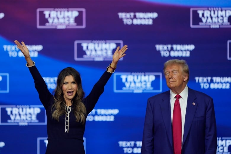 A dark haired woman raises her arms above her head while an older clean shaven man in a suit and tie stands nearby. Both are on a stage.