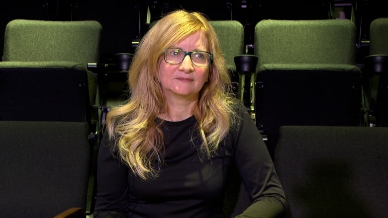 Woman with blond hair and wearing glasses. She has a serious expression and she's sitting in a theatre.