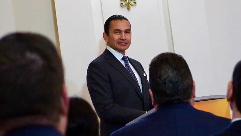 A man on a suit and a tie stands in front of a white wall.