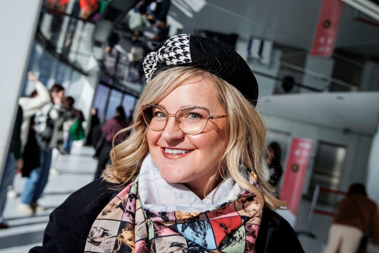 Fan smiles for the camera, wearing a houndstooth pattern beret and Taylor Swift Eras Tour scarf, with visitors behind her at the CN Tower's main observation deck.