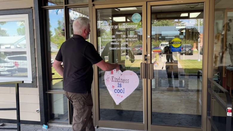 A man with grey hair, wearing slacks and a black golf T-shirt, opens a double glass door. The door has a pink heart on it that says 'Kindness is Cool' and a Pride flag sticker.