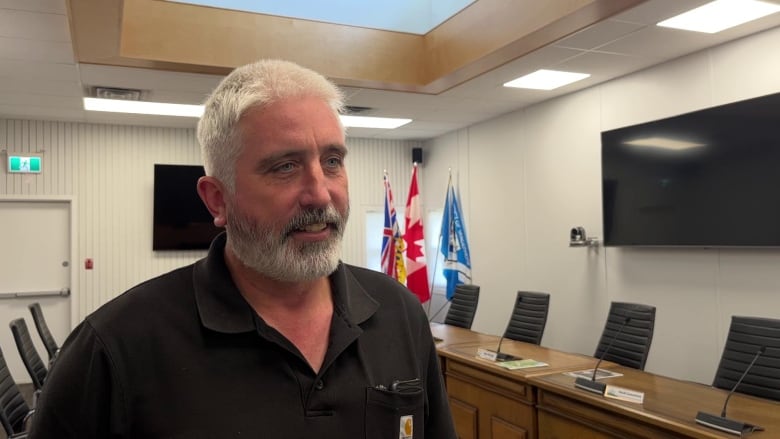 A man with grey hair and beard wearing a black golf T-shirt speaks in his board office.