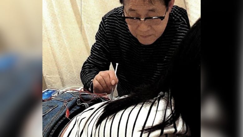 A man in glasses leans over a person's white top with a needle.