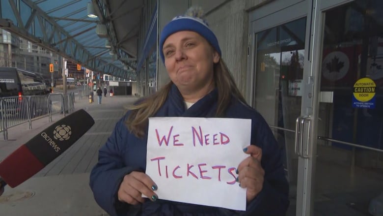 Photo of a woman outside holding a sign reading 'We need tickets.' 