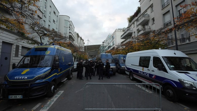 Police officers wearing black stand in the middle of a closed street. Blue police vans are also visible.