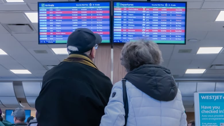 A couple stand looking at an airport arrival and departures board that shows a number of delayed and cancelled flights.