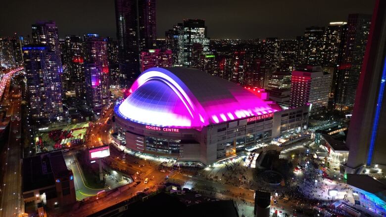 An aerial image shows Toronto's Rogers Centre lit in pink and purple hues at night as people gather for the Taylor Swift concert. 