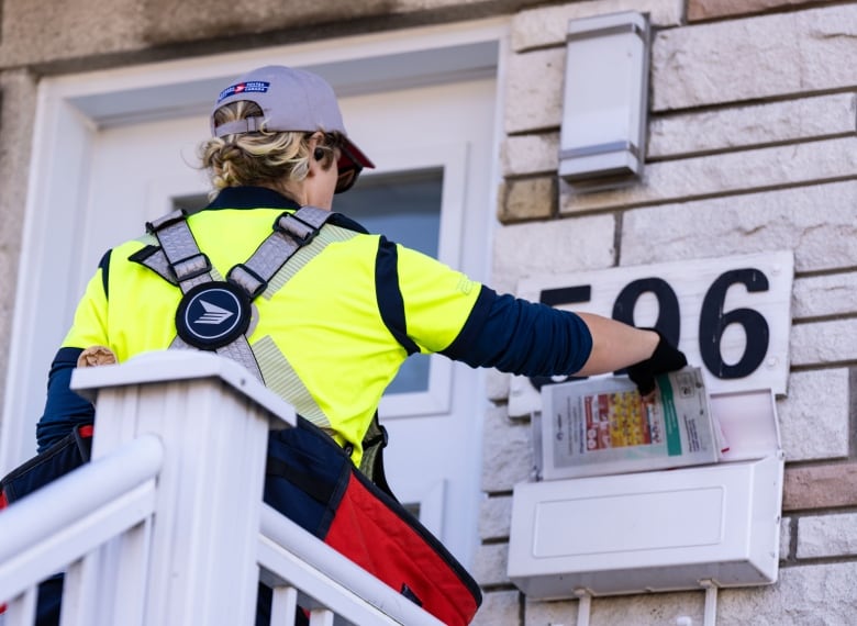 The back of a mail carrier who is wearing a baseball cap is shown. They are extending their right arm toward a mail box.