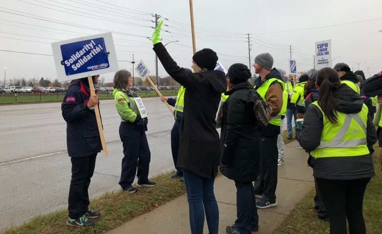 A group of people stand along the sidewalk wearing Canada Post jackets and holding signs. 