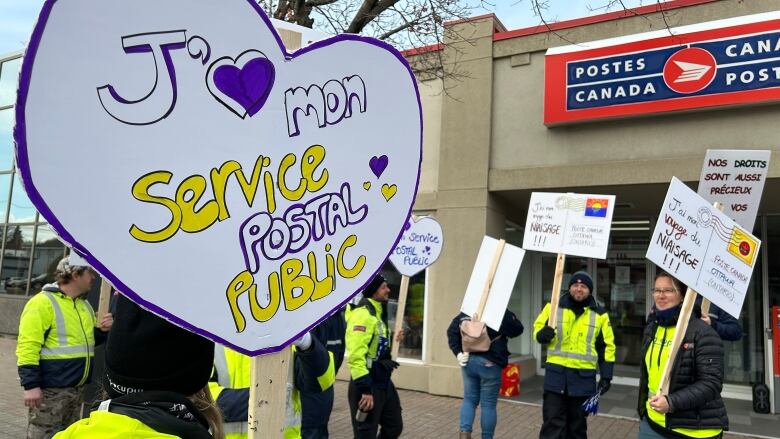 A scene outside a postal office where workers are striking. A heart-shaped sign reads 