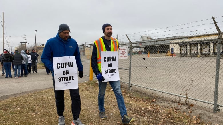 Canada Post workers wearing posters and walking the picket line, outside a fenced off facility.