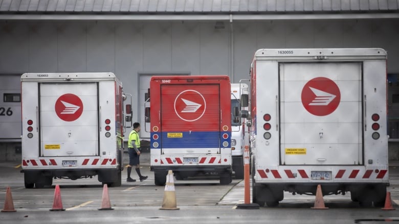 A worker in green high-viz clothing is seen walking between red-and-white delivery trucks at a mail sorting facility.