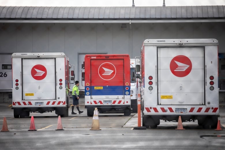 A worker in green high-viz clothing is seen walking between red-and-white delivery trucks at a mail sorting facility.