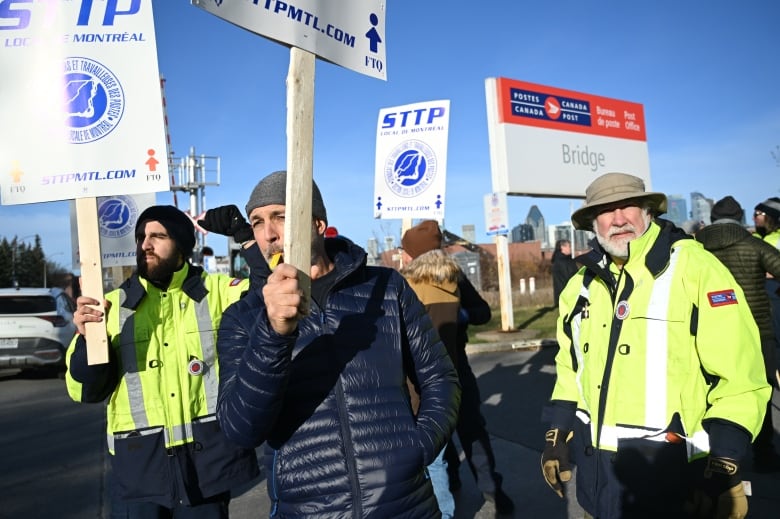 Workers bundled in winter jackets and toques picket outside a sorting plant on a clear day.