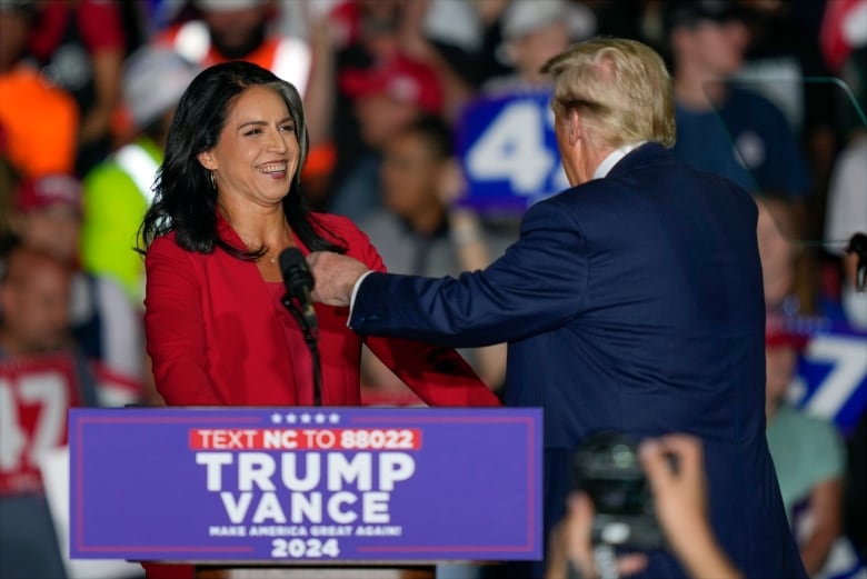 A dark-haired woman wearing a red suit jacket smiles from behind a podium as a man in a suit and tie approaches with his arm out.