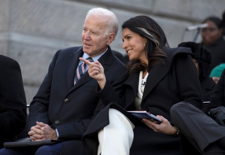 A white haired clean shaven man wearing a tie and a woman smile and are engaged in conversation in an outdoor photo, with both wearing winter coats.