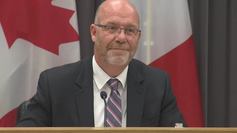 A smiling man in a suit in front of a Canadian and New Brunswick flag