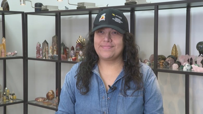 A woman in a jean shirt and ball cap and long, black hair sits in front of shelves containing crystals of all shapes and sizes.