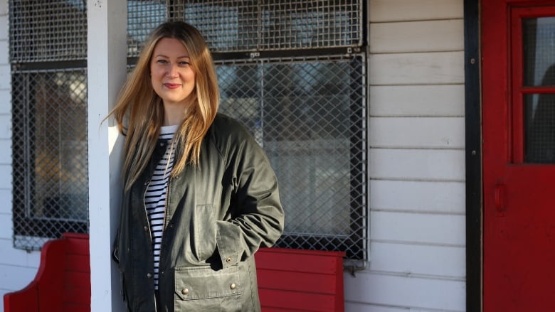 A white woman, with long amber hair, is smiling while leaning against a white wooden beam. She is wearing a dark green jacket, unzipped, revealing a black-and-white striped shirt. She is standing under the canopy of a white building with a red door and red bench.