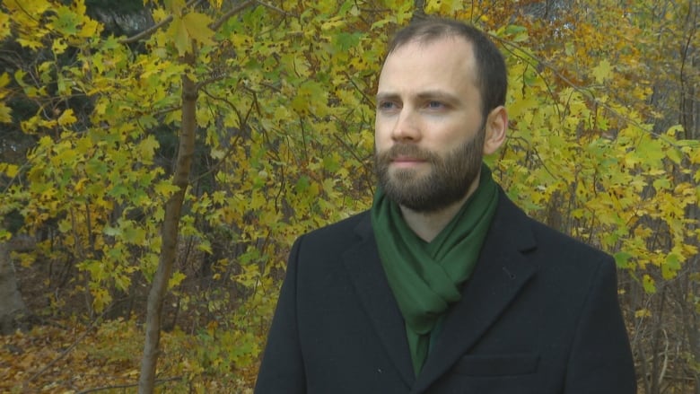 Anthony stands in front of trees with a black coat and green scarf on. 