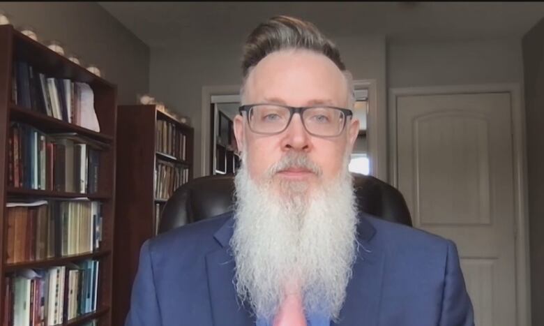 Man with long grey beard sitting beside bookshelf.