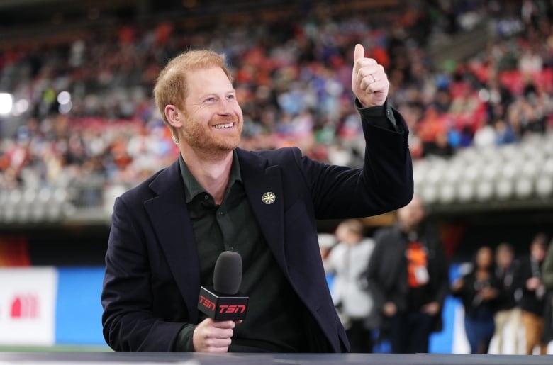 A man with red hair and a beard smiles while giving a thumbs up in the middle of a football stadium.