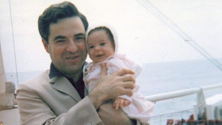 A clean shaven dark haired man holds a baby in a bonnet in an outdoor photo that appears decades old.