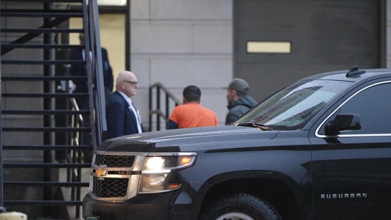 three men stand outside a courthouse, one is wearing orange and has his back turned to the camera