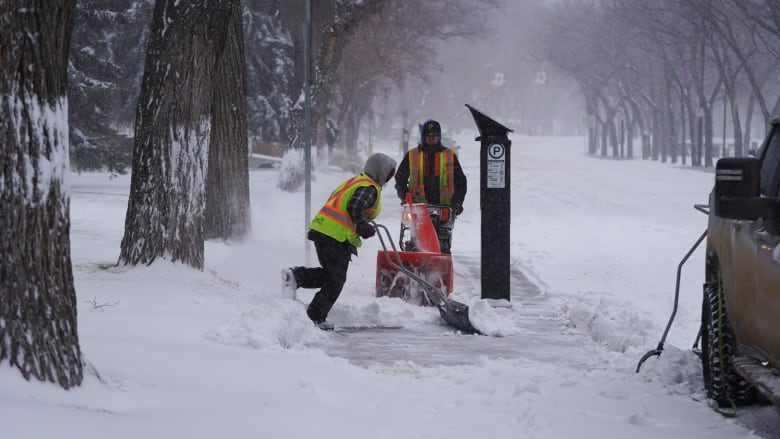 Two people in neon vests work to clear a sidewalk, one with a shovel and one with a snowblower.