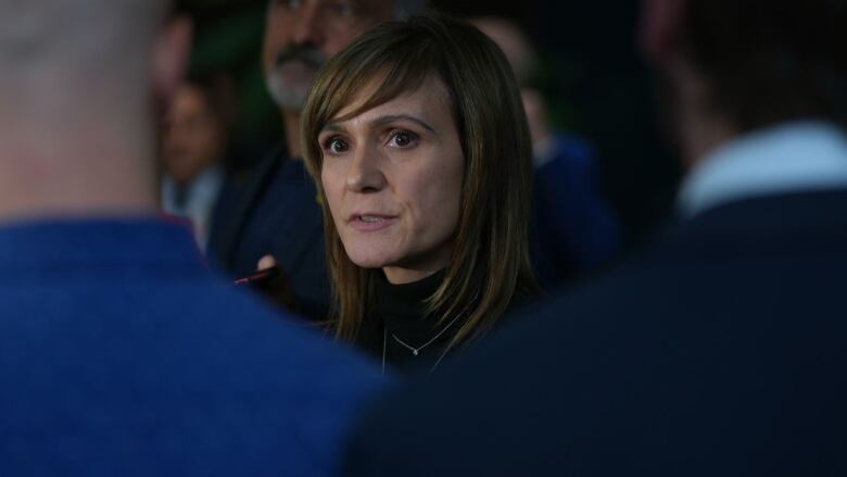 A woman is pictured in a close-up image. she stands in a crowd of people while speaking, with blurry shoulders in the image's foreground.