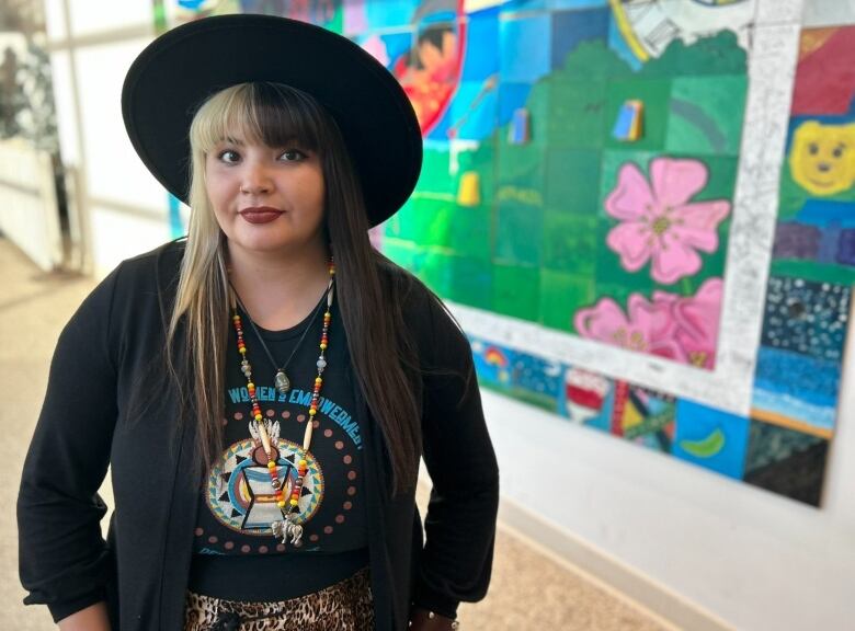 A woman with black and blonde hair stands in an art gallery setting. She is wearing a bead necklace and a black rimmed hat. 