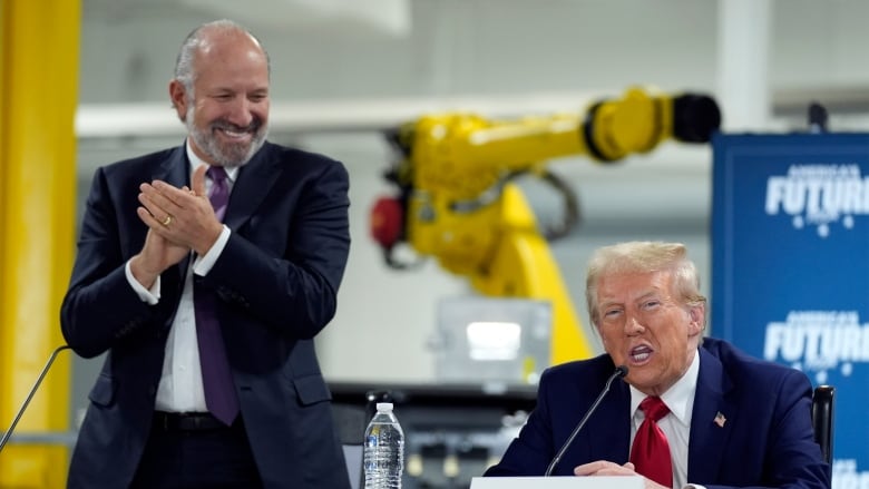 A smiling man in a suit stands and claps next to a seated Donald Trump who is speaking into a microphone. 