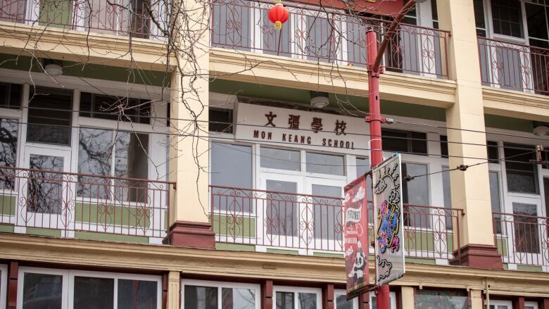 A three-level tan structure is draped with red Chinese banting with the words Mon Keang School in Chinese lettering over a second-story entrance.