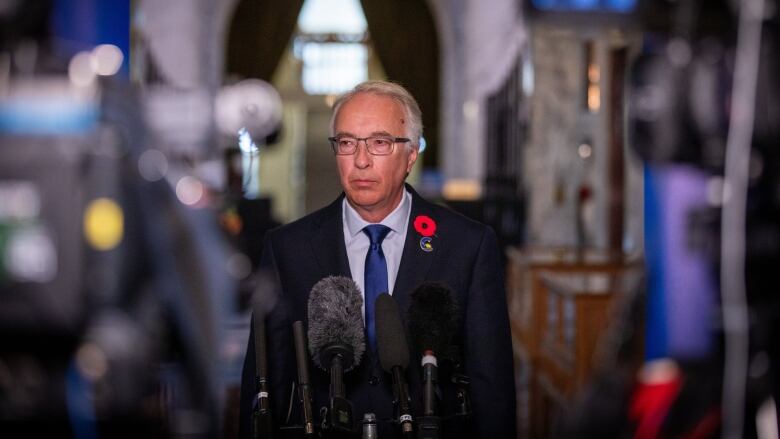 A man looks to his right as he stands in front of a cluster of microphones.