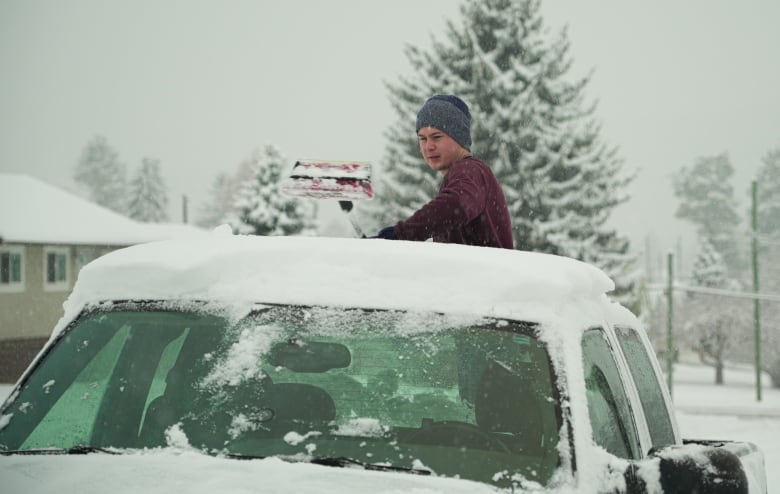 A person in a toque clears snow from the roof of a vehicle.