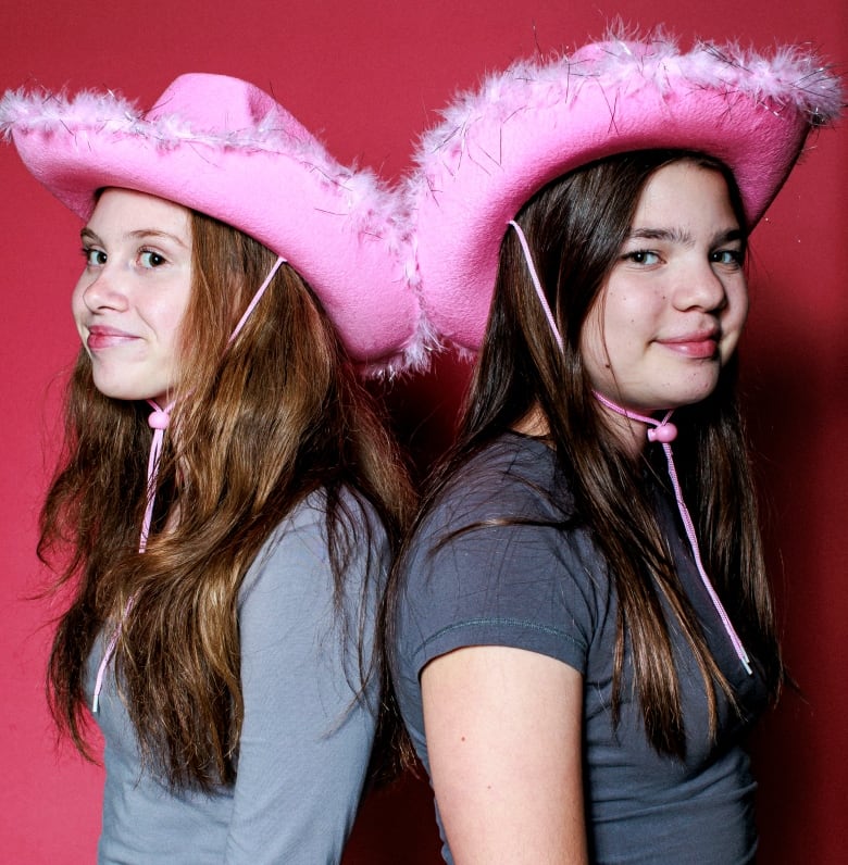 Two young girls in pink cowboy hats standing with their backs to one another and looking into the camera for a portrait.