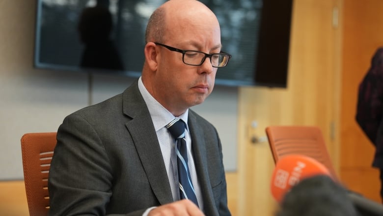 A man in a suit sits at a table before a microphone.