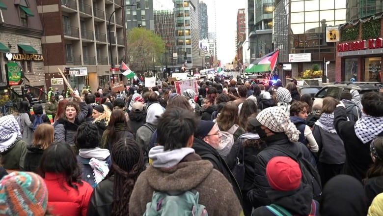 a group of students with keffiyeh scarves and pro-Palestinian signs outside a beige building.