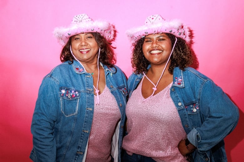 Smiling, two women stand against a pink background wearing matching pink cowboy hats, pink metallic sweaters and jean jackets.