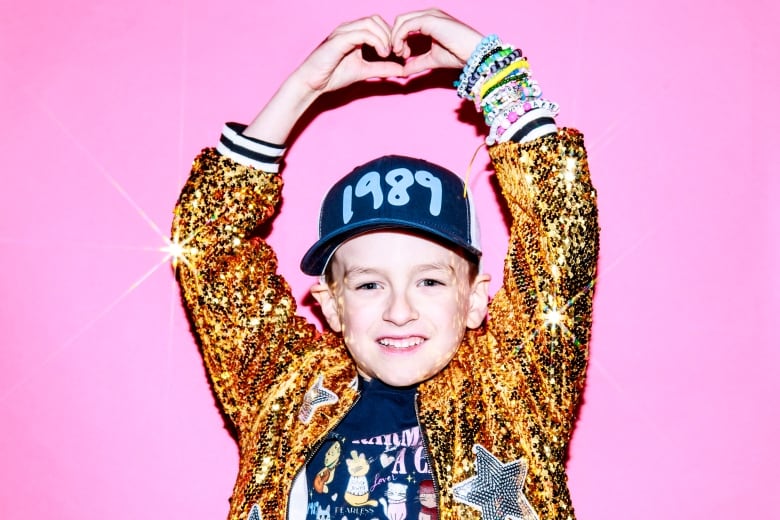 A young child makes a heart-shape with their hands wearing a gold sequin bomber style jacket and baseball hat that reads: 1989.