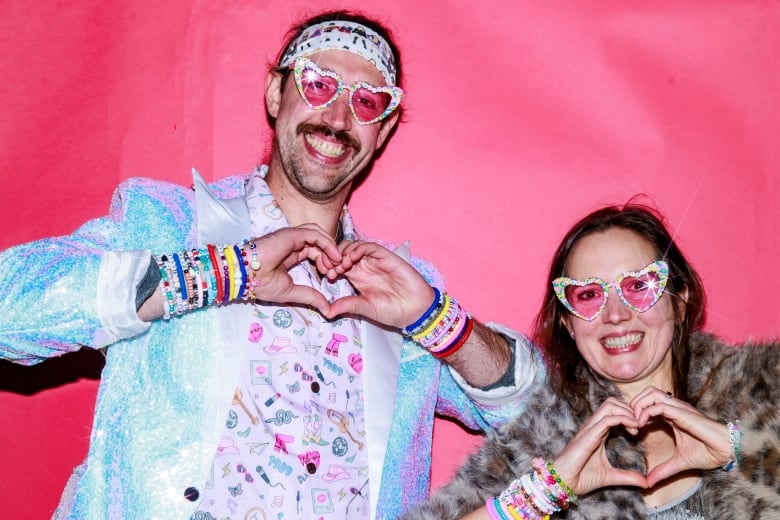 A man and woman make heart-shapes with their hands, wearing Taylor Swift-themed outfits and smiling at the camera.