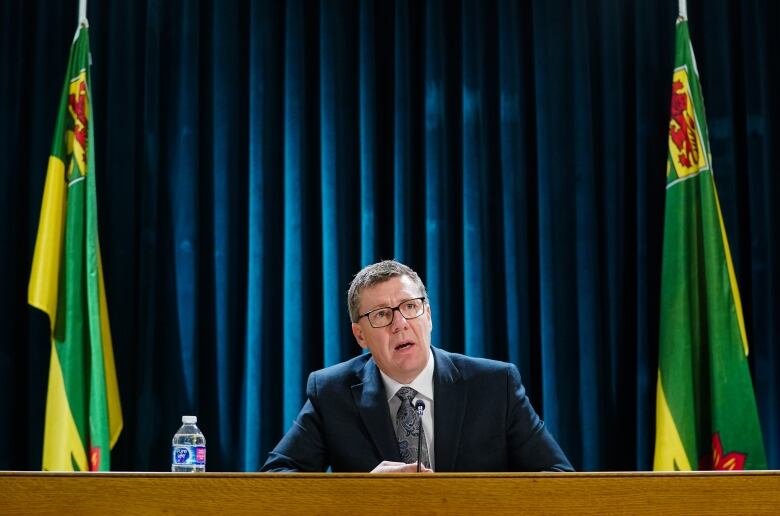 A man ina  suit and glasses sits at a desk speaking into a microphone.